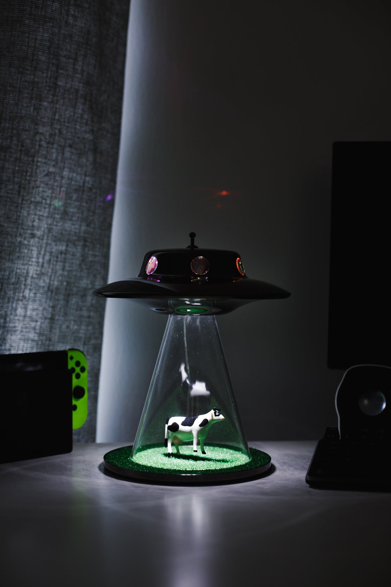 A UFO themed bedroom desktop alien lamp pictured next to a Nintendo Switch. This UFO lamp is the ultimate bedroom accessory and gift for alien lovers and outer space fanatics. Get your alien abduction lamp today!